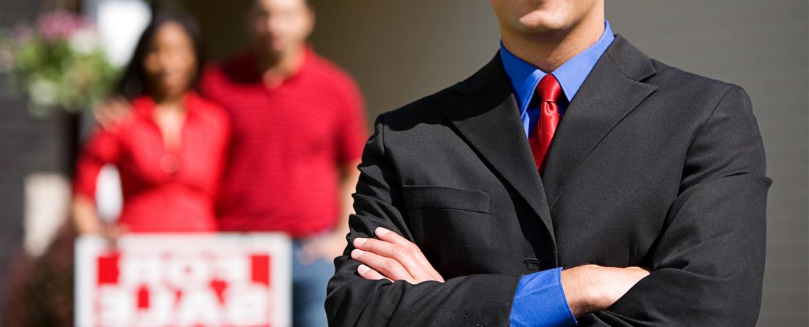 White male REALTOR in the foreground with sellers in background in front of for sale sign